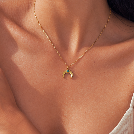 A close-up lifestyle shot of a model wearing a delicate gold chain necklace with a crescent moon pendant featuring a small turquoise stone.