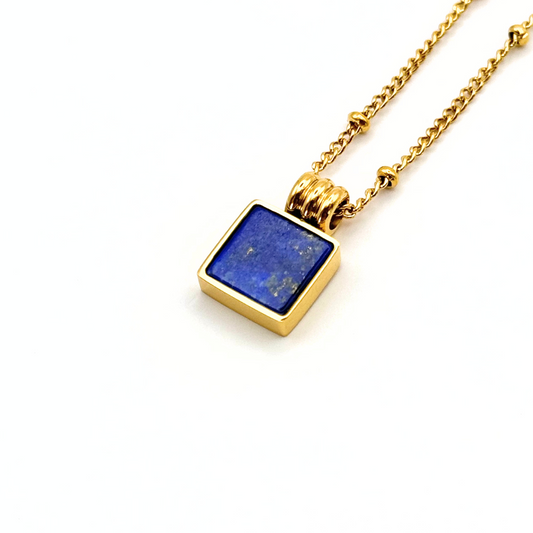 A gold chain necklace with a square pendant featuring a deep blue lapis lazuli stone in a gold bezel setting, shown on a clean white background.