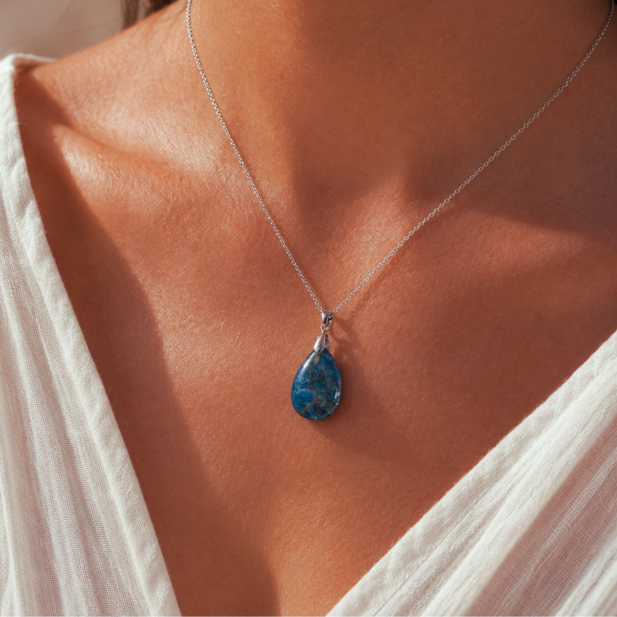 A close-up of a woman's neck and chest wearing a delicate silver chain necklace with a teardrop-shaped Apatite stone pendant.