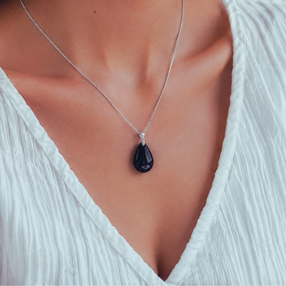 A close-up of a model wearing a delicate silver steel necklace with a polished black teardrop black obsidian pendant, styled with a white V-neck top.