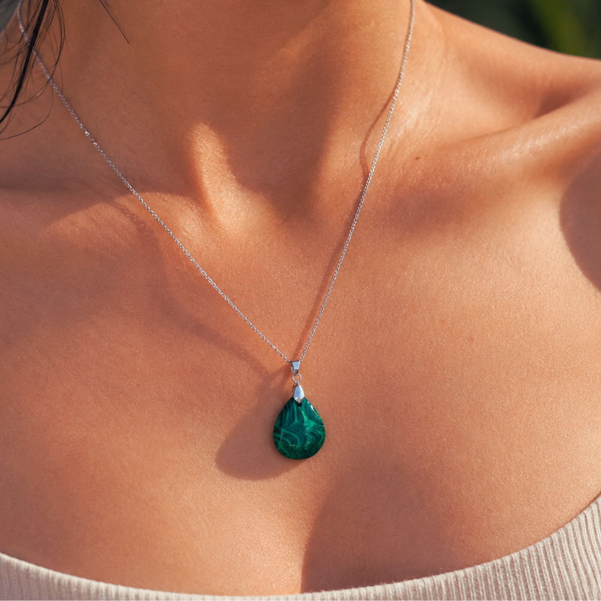 A close-up of a woman's neck and chest wearing a delicate silver chain necklace with a teardrop-shaped Malachite pendant.
