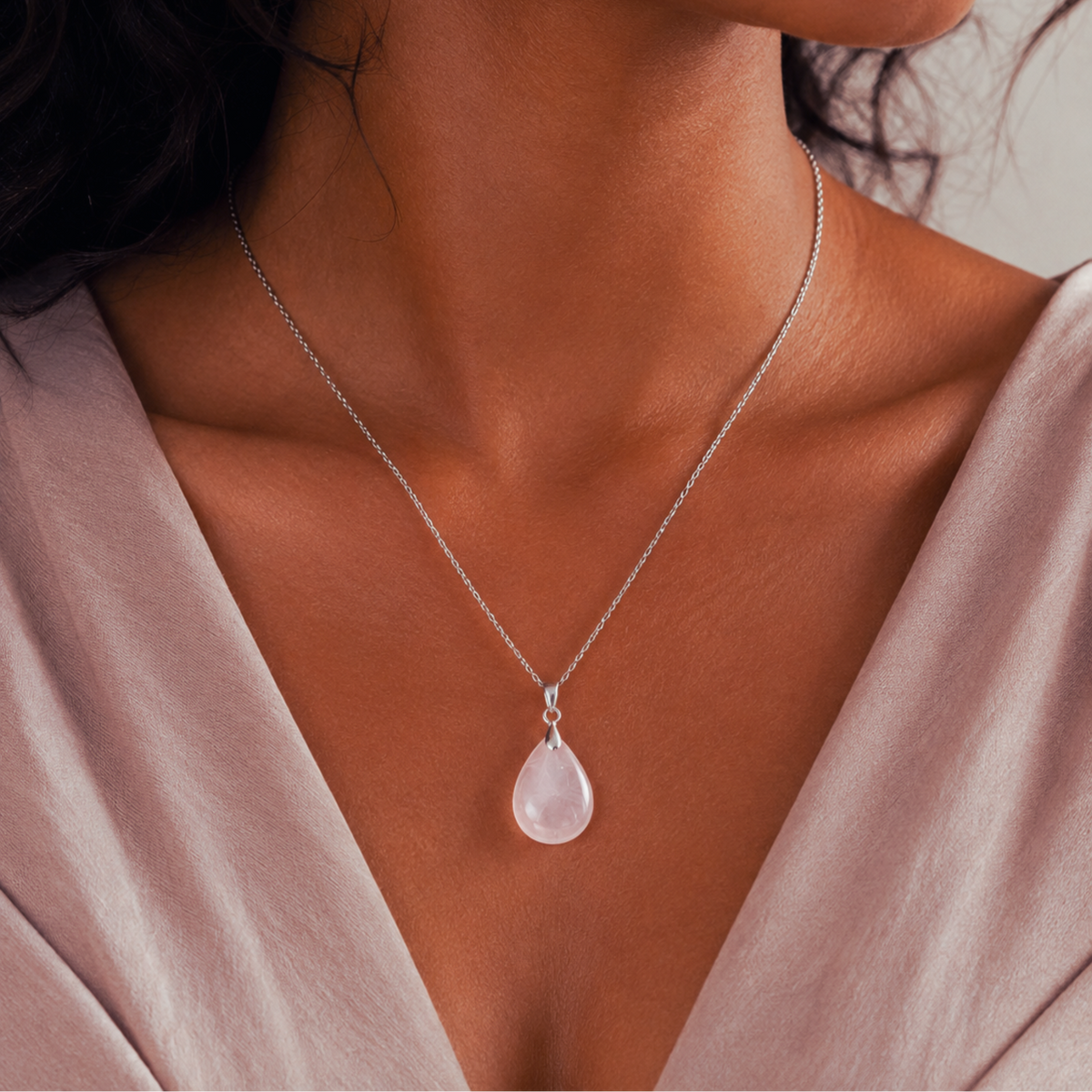 A close-up of a woman wearing a delicate silver chain necklace with a teardrop-shaped rock quartz stone pendant, paired with a soft mauve top.