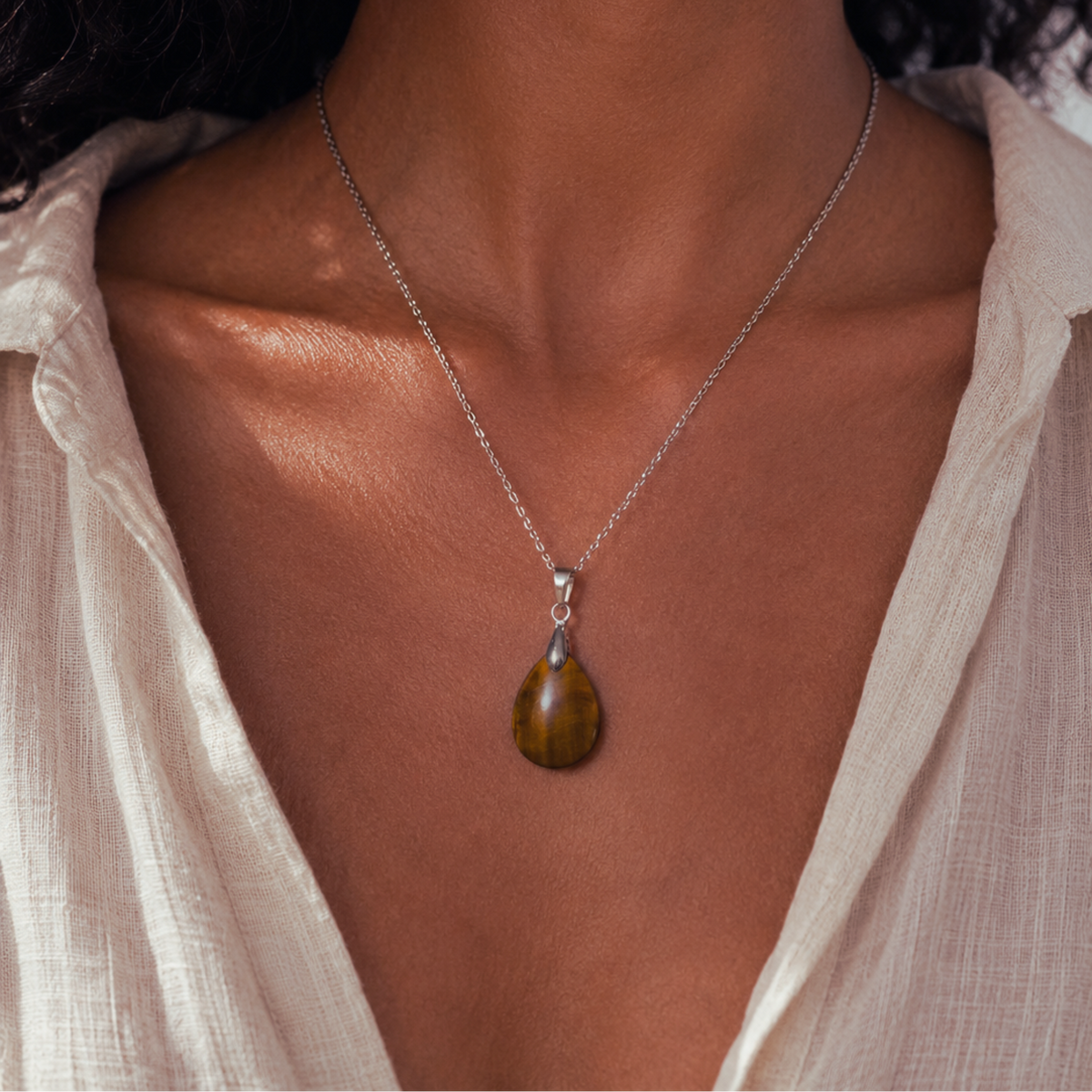 A close-up of a model wearing a silver chain necklace with a teardrop-shaped brown tiger's eye pendant, styled with a light beige linen top.