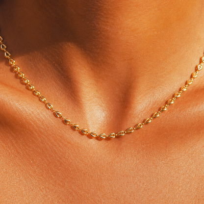 A close-up of a delicate gold chain necklace worn around the neck of a model with warm, sunlit skin.