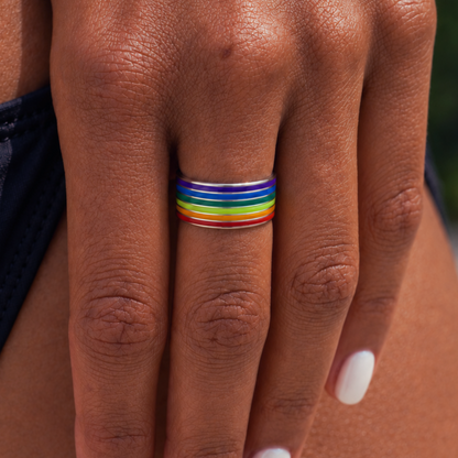 A close-up of a person's hand wearing a thin silver band ring with multiple horizontal rainbow-colored stripes, including red, orange, yellow, green, blue, and purple.